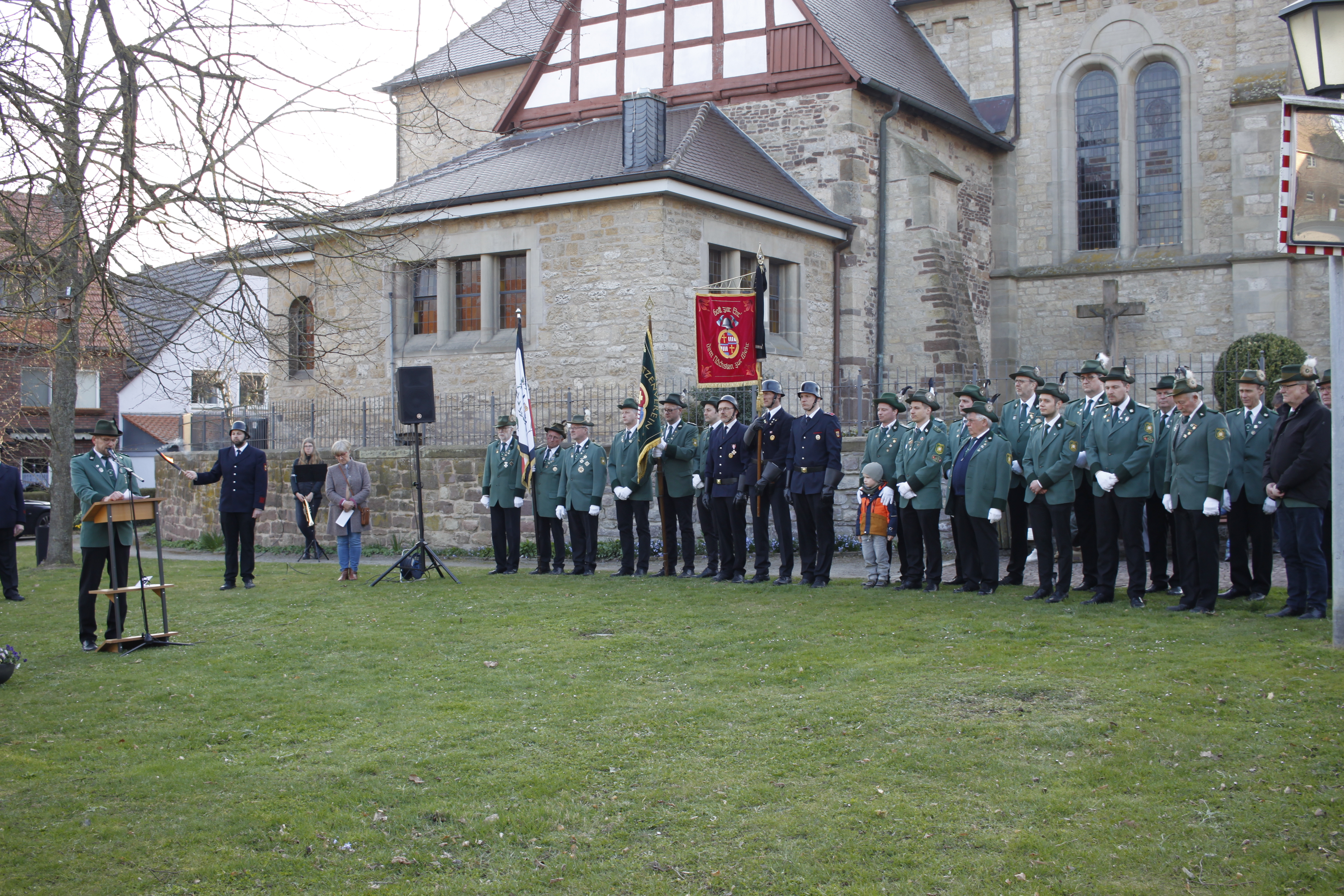 Schützen und Feuerwehrleute in Uniform stehen mit ihren Fahnen auf einer Wiese vor einer Kirche
