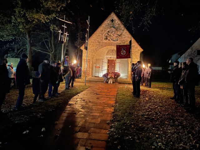 Im Dunkeln erleuchtete Kapelle vor der ein Kranz niedergelegt wurde, Menschen stehen davor.