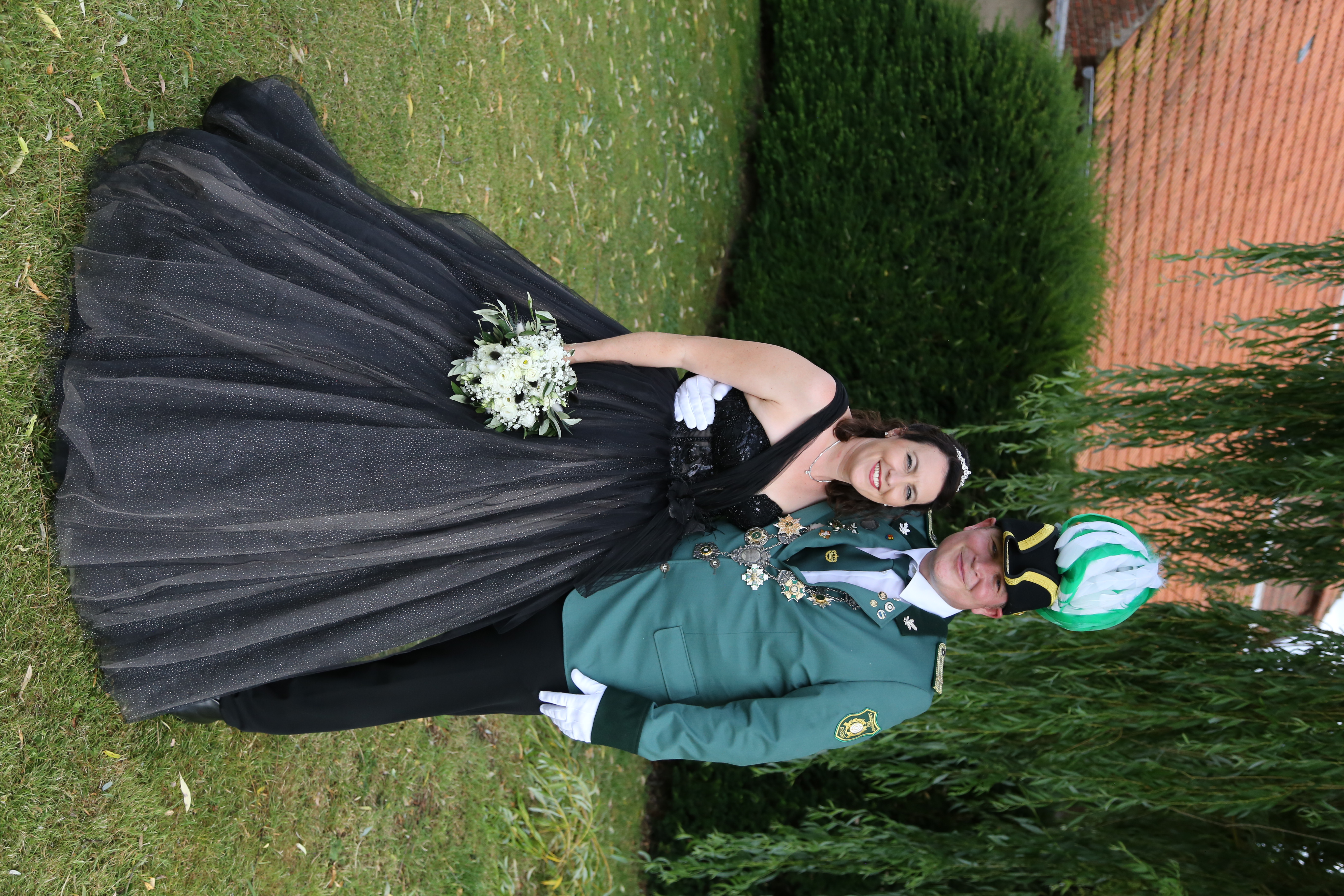 Eine Frau im Festkleid und ein Mann in grüner Uniform mit Hut mit Federbusch und Königskette stehen unter einer Trauerweide auf dem Rasen und halten sich im Arm